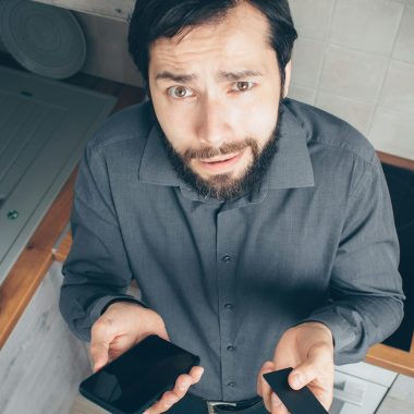 A confused man indoors looks up while holding a smartphone and credit card, concerned about a financial issue.